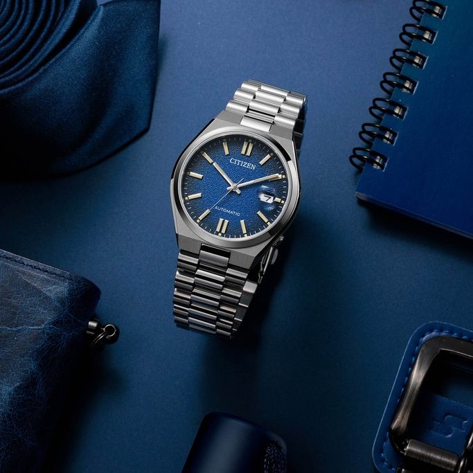 Eine elegante Citizen-Uhr mit blauem Zifferblatt und silbernem Armband wird zusammen mit blauen Accessoires wie Krawatte, Brieftasche und Notizbuch auf einer passenden blauen Fläche präsentiert.
