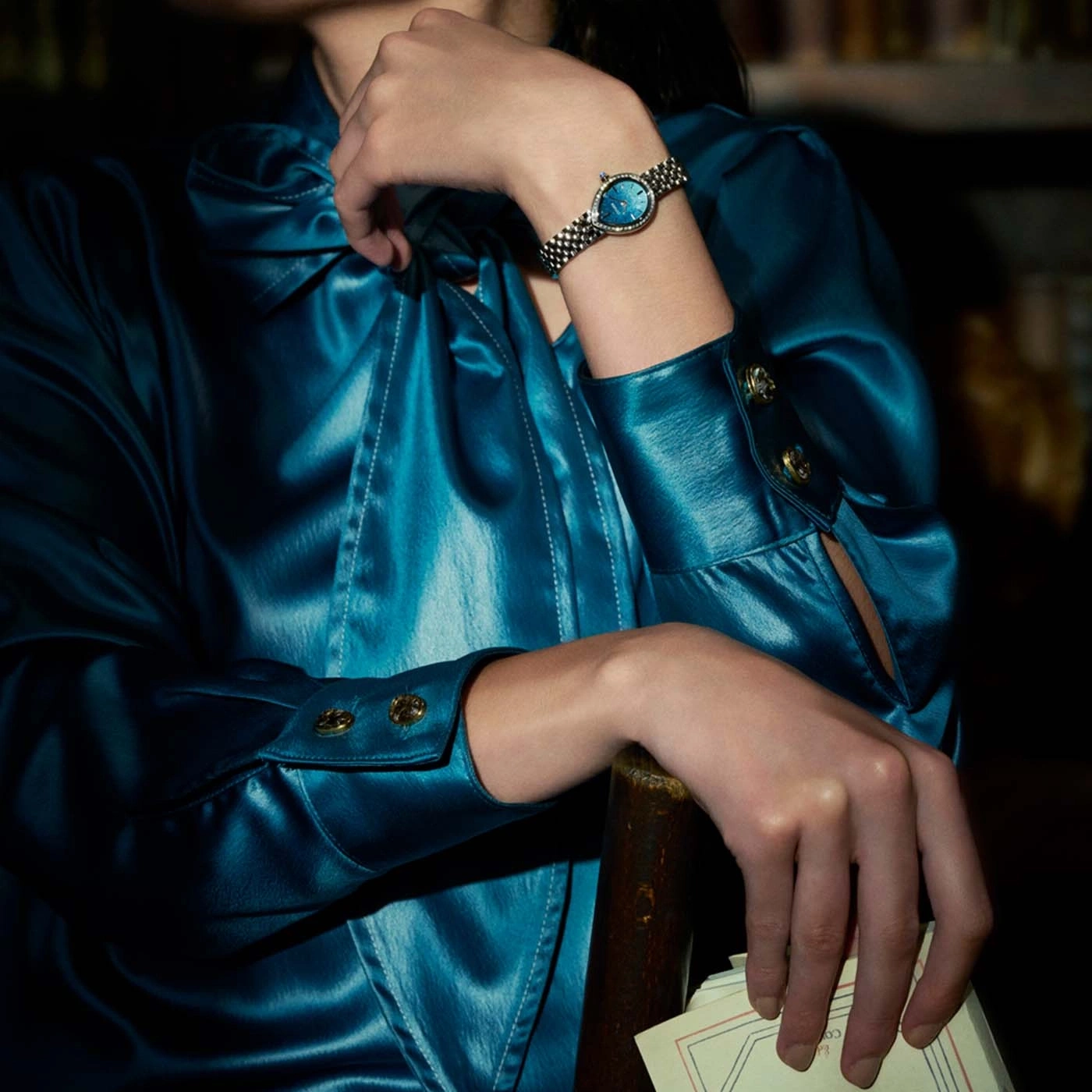 Frau in blauem Satinkleid mit Uhr und Papieren.