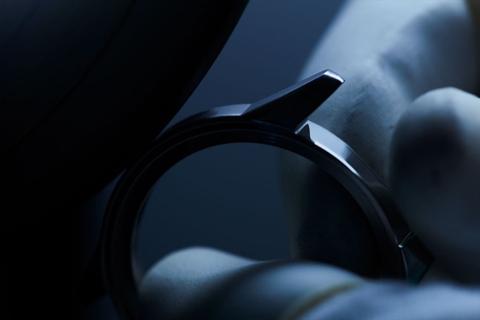 Nahaufnahme einer Uhrenkomponente, gehalten von einer behandschuhten Hand, mit dunklem Hintergrund.