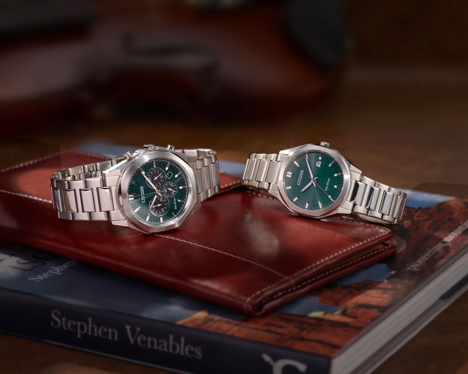 Zwei silberne Citizen-Uhren mit grünen Ziffernblättern liegen auf einer braunen Brieftasche über einem Buch und bilden ein elegantes, detailreiches Stillleben.
