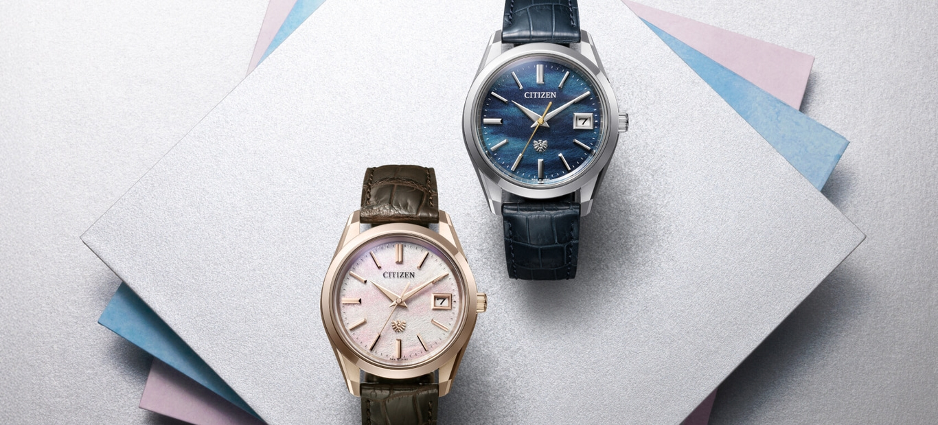 This image displays two Citizen watches: a blue dial watch with a dark blue strap, and a light dial watch with a brown strap, both on textured, layered surfaces.