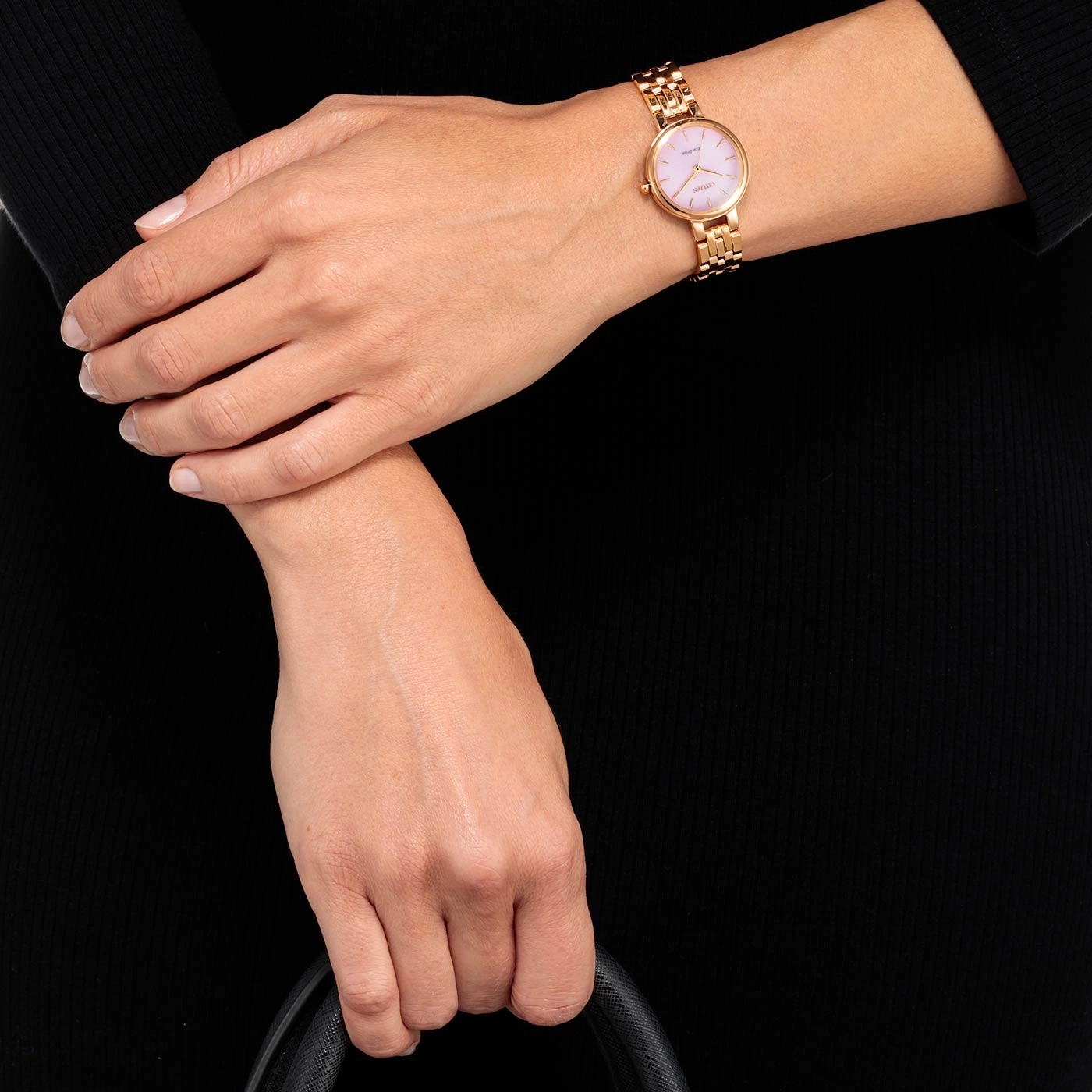 Frau mit goldener Uhr und rosa Zifferblatt, hält eine schwarze Tasche.