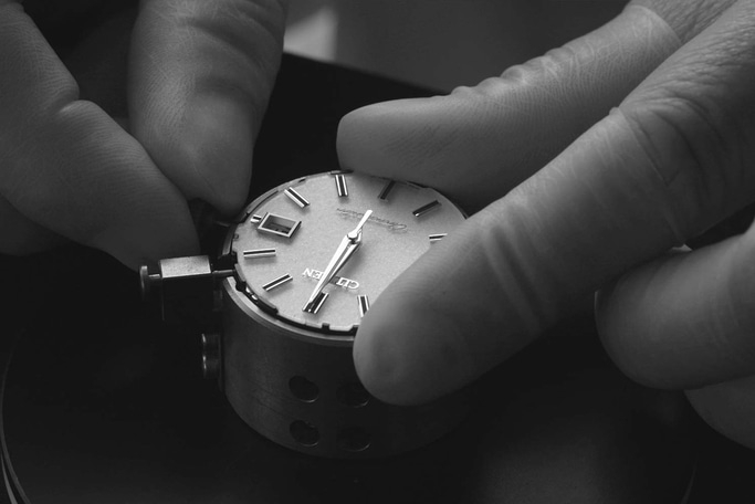 This black and white close-up shows a watchmaker's hands precisely working on a watch movement and dial. The hands are positioning or adjusting components, emphasizing the intricate and delicate craftsmanship.