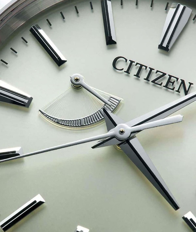 This close-up image features a Citizen watch dial with a subtle off-white color. It showcases sleek, polished hour markers and hands, along with a unique power reserve indicator. The "CITIZEN" logo is prominently displayed.