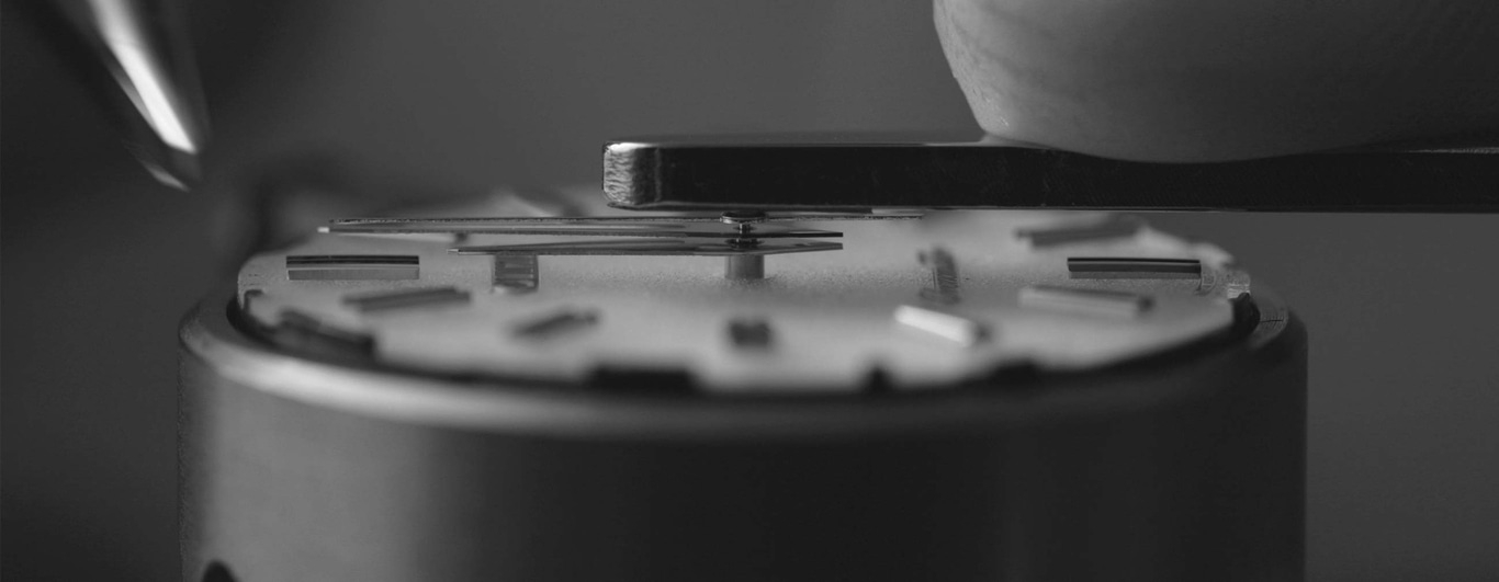 This black and white close-up shows watchmaker's tools precisely working on a watch dial. An instrument positions a component over minute markers, highlighting the delicate, precise nature of watch assembly.