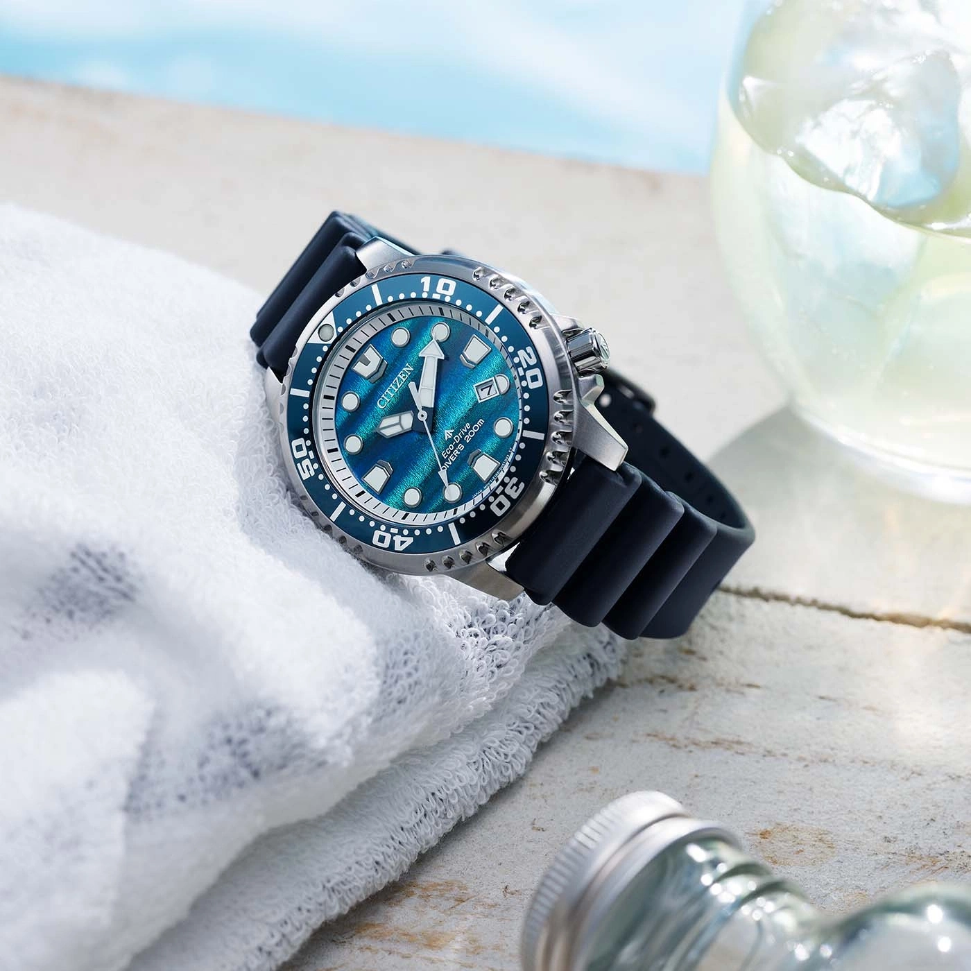 Eine blaue Citizen Eco-Drive Taucheruhr liegt auf einem weißen Handtuch neben einem Glas mit Eisgetränk.