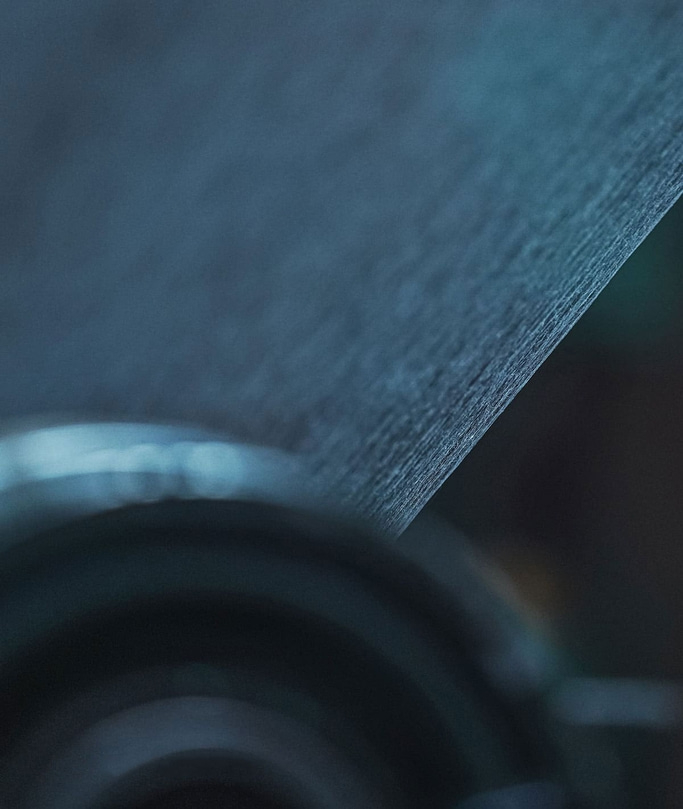 This close-up abstract image, in cool blue tones, features a textured surface, possibly fabric or paper, diagonally across the frame. It appears to be moving over a blurred, dark circular object, suggesting a winding or industrial process.