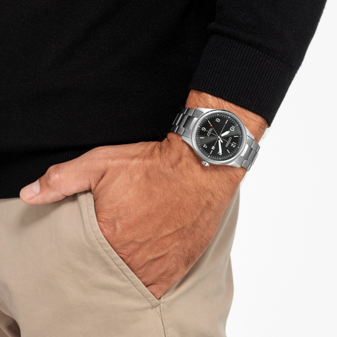 Mann mit Uhr, Hand in der Tasche, schwarzer Pullover, beige Hose, weißer Hintergrund.