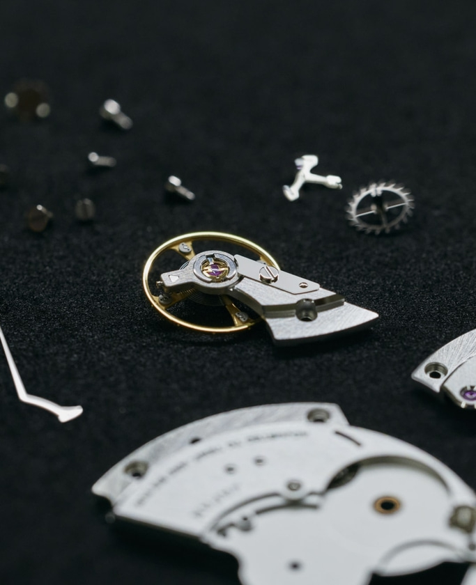 This close-up features an array of intricate watch components scattered on a dark, textured surface. Gears, screws, and parts of the movement are visible, highlighting the delicate precision and numerous elements involved in watchmaking.