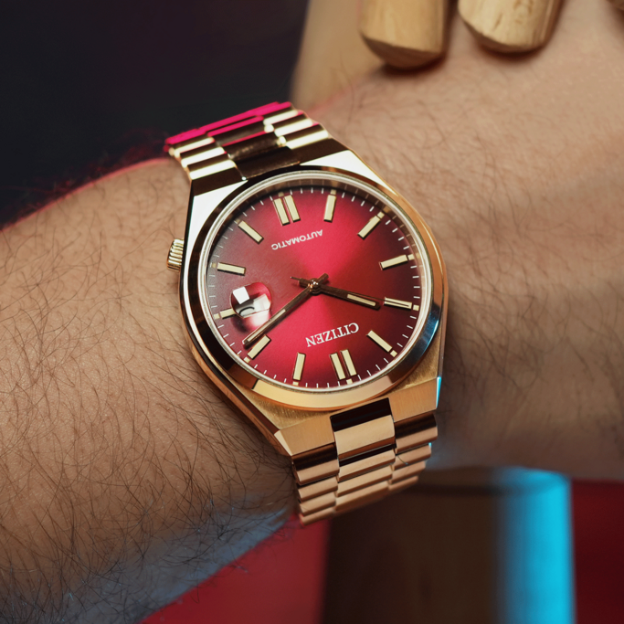 Nahaufnahme einer goldenen Citizen-Uhr mit rotem Zifferblatt, die an einem behaarten Handgelenk getragen wird. Die Uhr hat ein goldenes Armband und ist scharf gestellt, mit einem unscharfen Hintergrund aus Haut und einem Holzobjekt.
