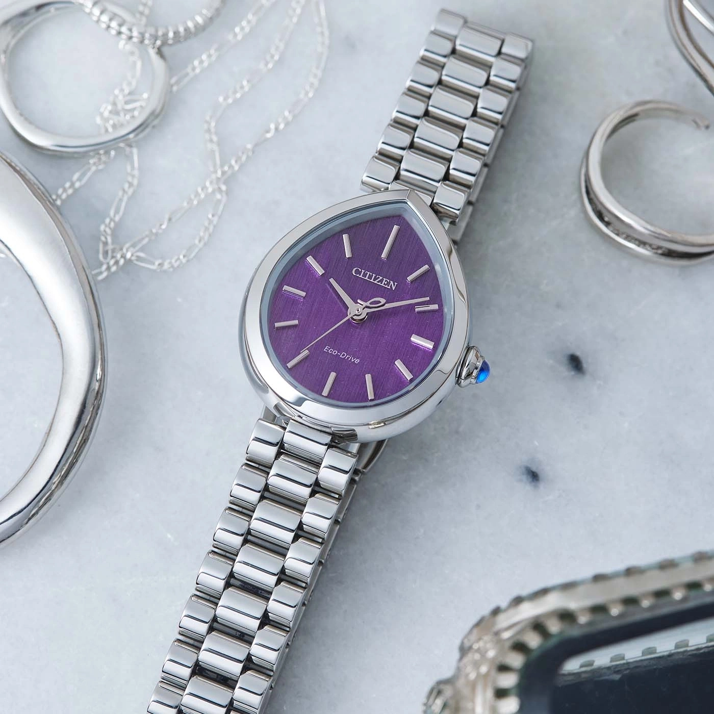 Silberne Uhr mit violettem Zifferblatt und silbernem Armband, umgeben von Schmuck.