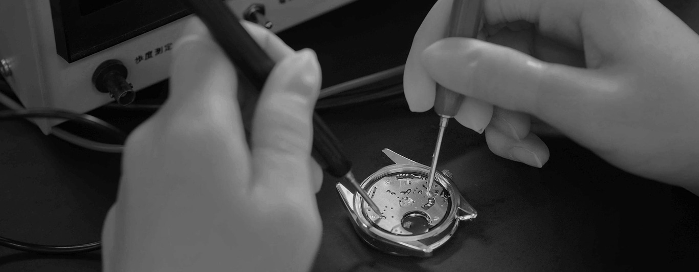 This black and white image shows a watchmaker's hands using tools to meticulously work on a watch movement. The intricate components of the open watch case are visible, highlighting the precision and skill involved.