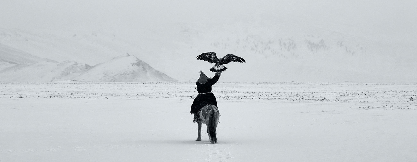 This black and white image shows a lone Kazakh eagle hunter on horseback in a vast, snow-covered landscape. The hunter holds a large eagle aloft. Distant mountains and sparse trees complete the desolate, yet majestic scene, capturing a timeless tradition