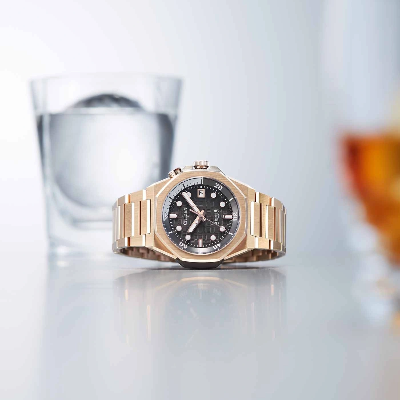 Goldene Uhr mit schwarzem Zifferblatt, Glas Wasser und reflektierender Oberfläche.