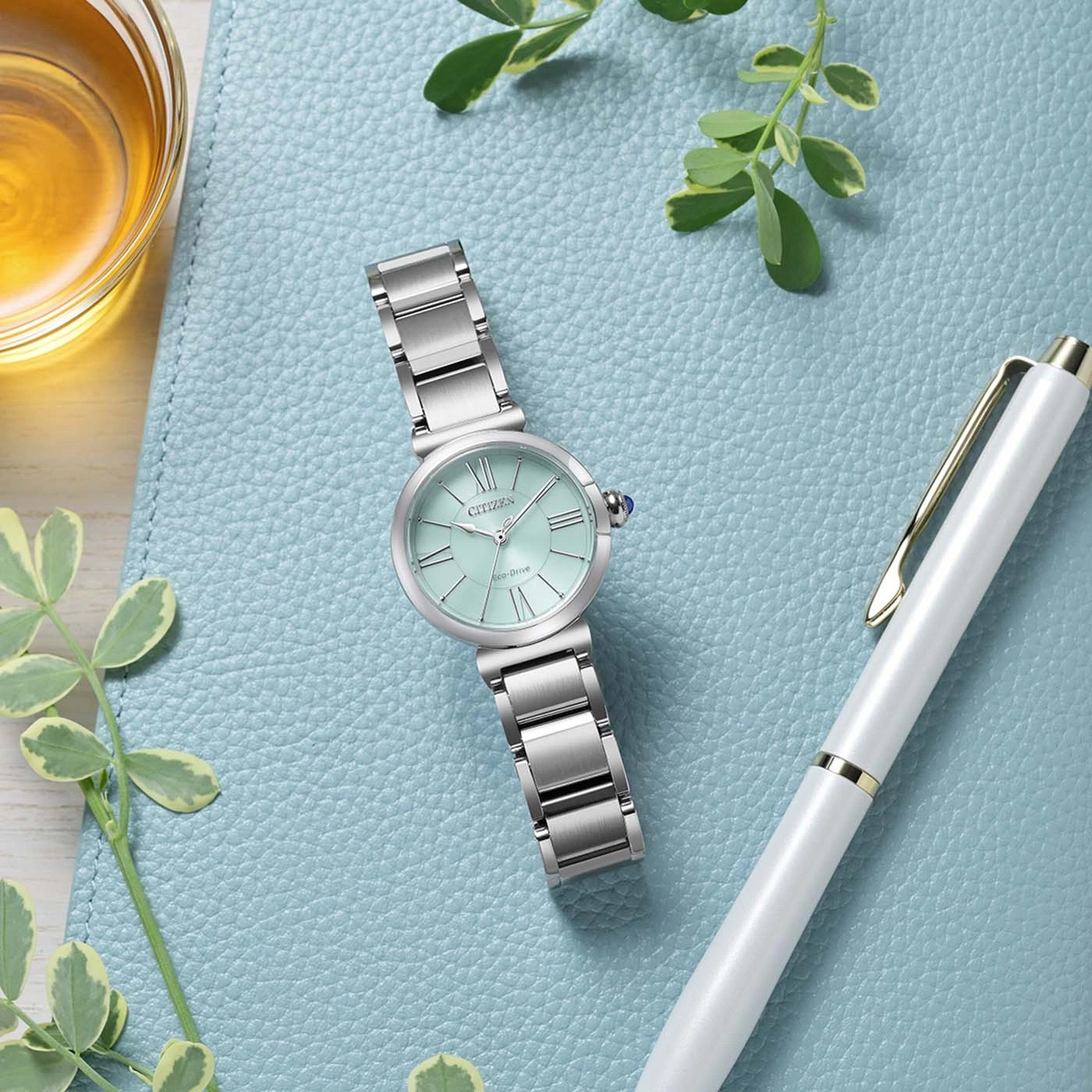 Eine mintgrüne Citizen Eco-Drive Uhr mit silbernem Band liegt auf einer blauen strukturierten Oberfläche neben einem weißen Stift und einem Glas mit bernsteinfarbener Flüssigkeit.