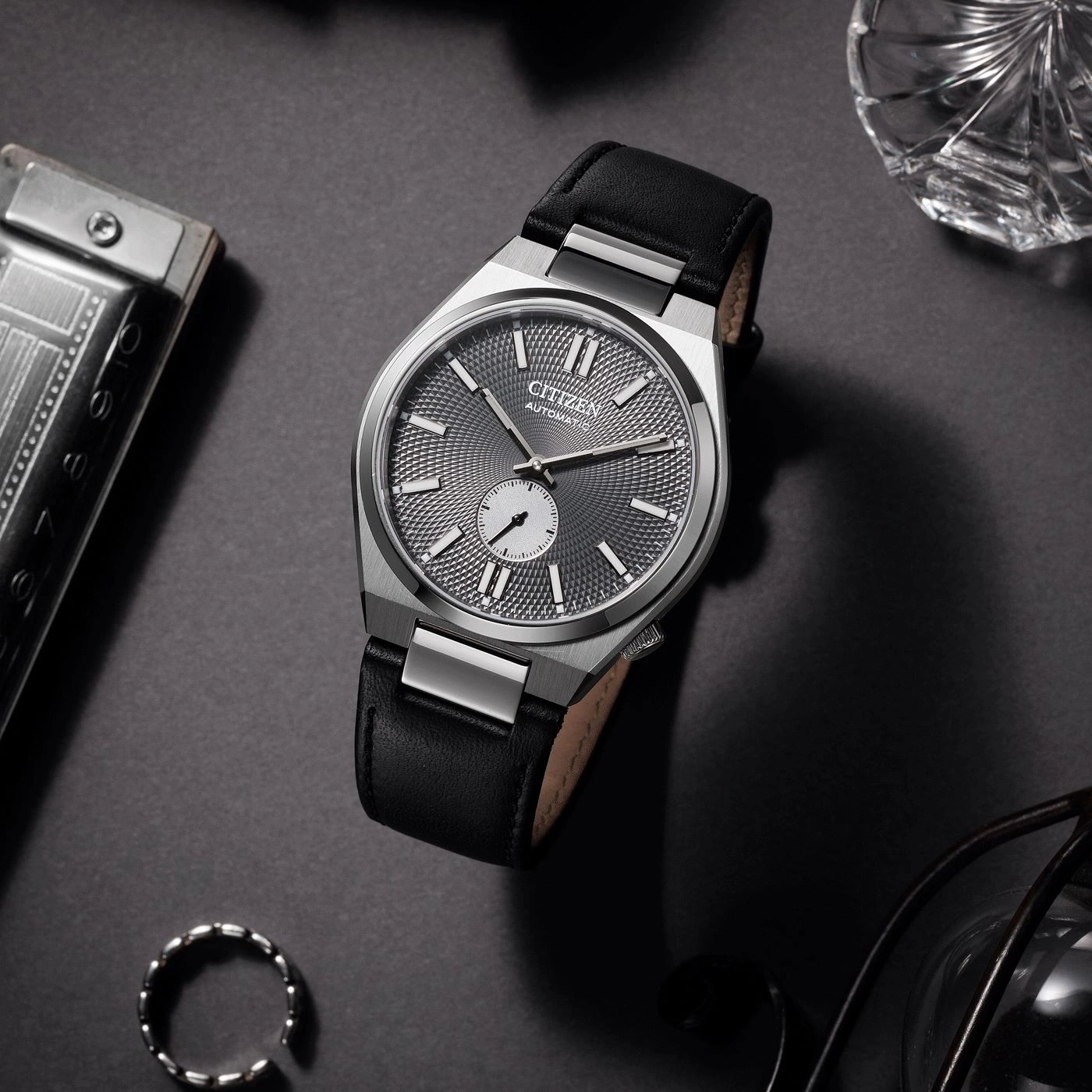 A stylish Citizen automatic watch with a black leather strap rests on a dark surface, surrounded by a harmonica, a crystal glass, a ring, and eyeglasses.
