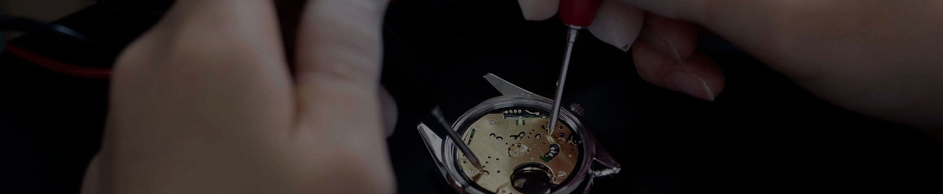 This close-up image shows a watchmaker's hands, one gloved, meticulously working on an open watch case with tools. The intricate internal components of the watch movement are visible, highlighting the delicate precision required for repair or assembly.