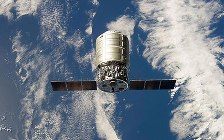 The image shows a space satellite orbiting Earth with a backdrop of clouds and blue sky.