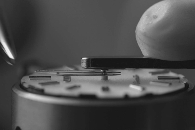 This black and white close-up shows watchmaker's tools precisely working on a watch dial. An instrument positions a component over minute markers, highlighting the delicate, precise nature of watch assembly.
