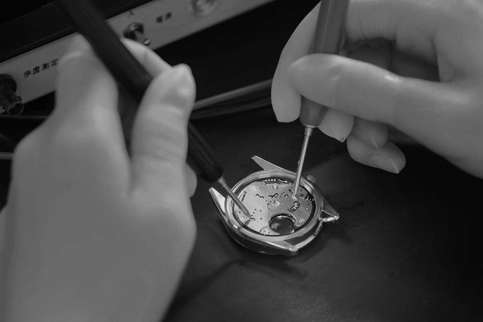 This black and white image shows a watchmaker's hands using tools to meticulously work on a watch movement. The intricate components of the open watch case are visible, highlighting the precision and skill involved.