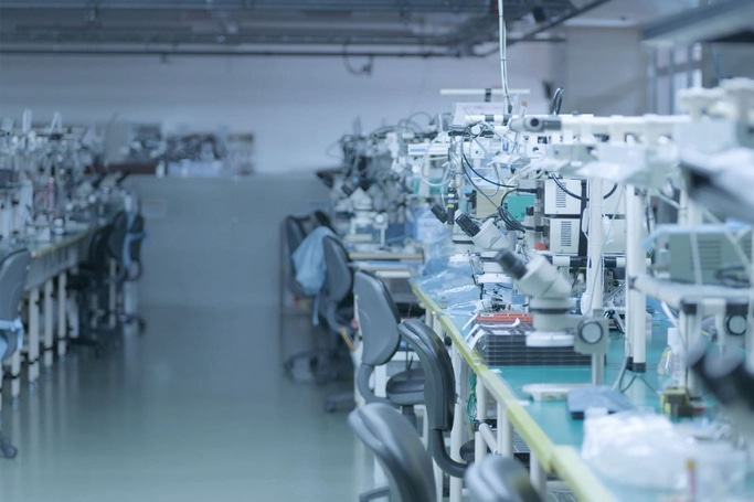 This image depicts a clean, organized factory floor or laboratory with rows of workstations. Each station features precision equipment, likely microscopes and tools, suggesting an environment focused on electronics assembly or intricate manufacturing.