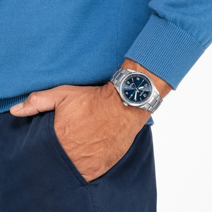 Mann mit Uhr, blauem Pullover und marineblauer Hose mit Hand in der Tasche.