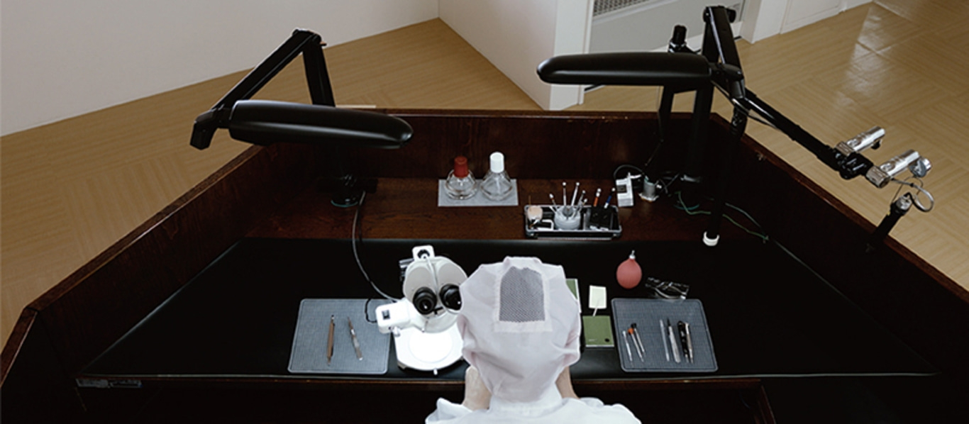 Eine Person mit Haarnetz arbeitet an einer Werkbank mit Mikroskop und Werkzeugen.