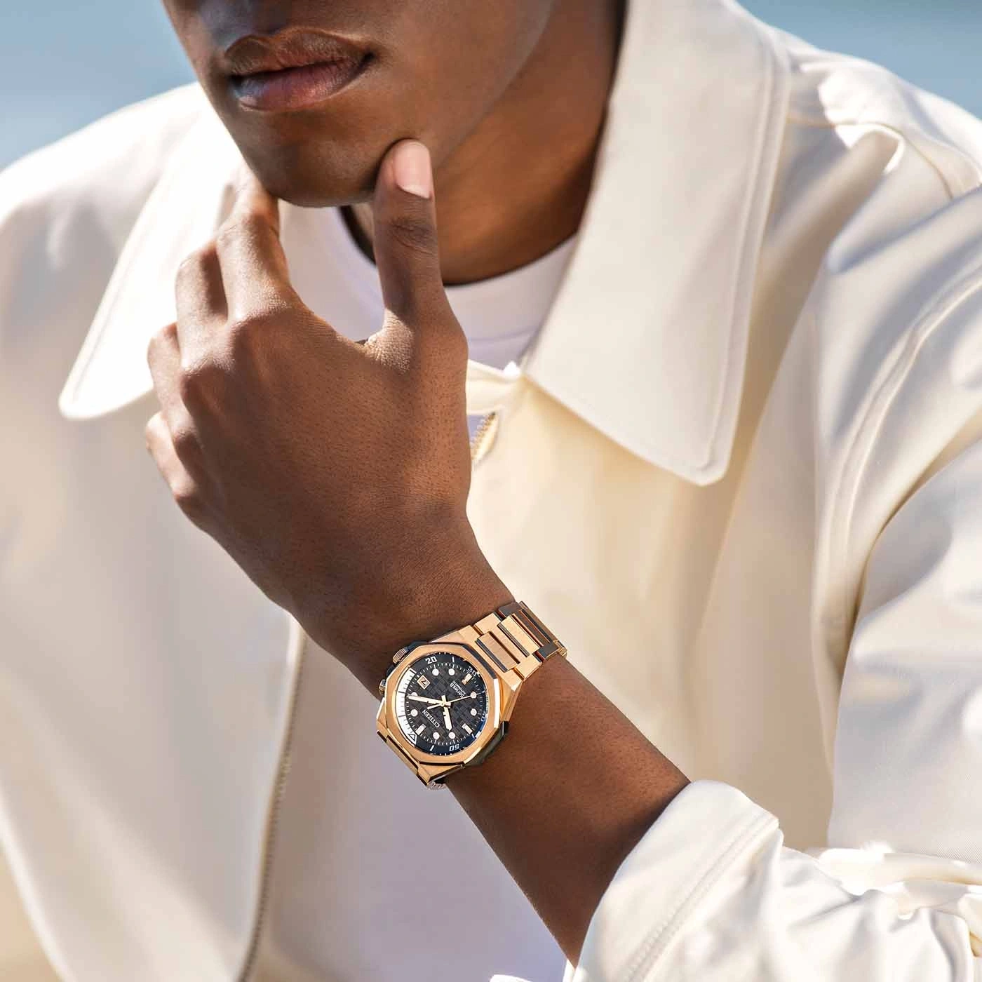 Mann mit goldener Uhr, posiert mit der Hand am Kinn, stilvoll gekleidet.