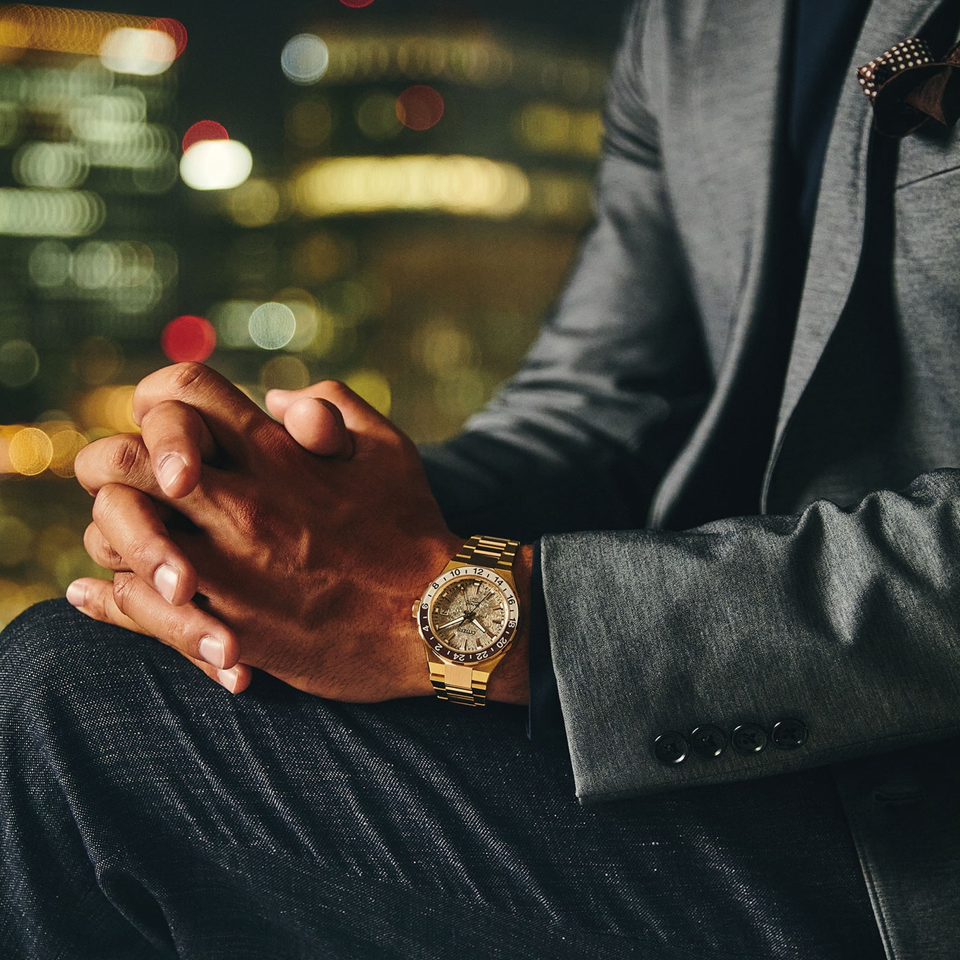 Mann im Anzug mit goldener Uhr, Hände gefaltet, Stadtlichter im Hintergrund.