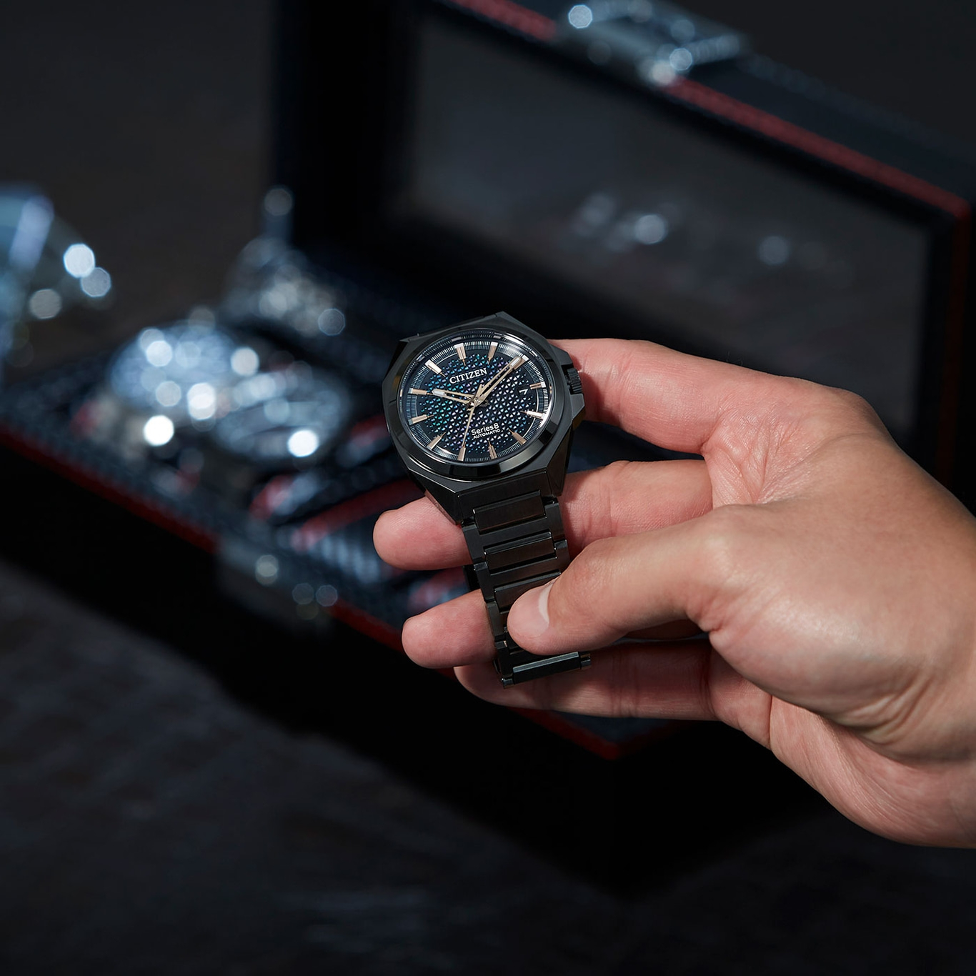 Eine Person hält eine schwarze Citizen-Uhr vor dunklem Hintergrund.