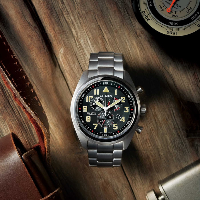 A Citizen Eco-Drive Titanium watch rests on a wooden surface, accompanied by a brown leather bag and an altimeter, creating a sense of adventure.
