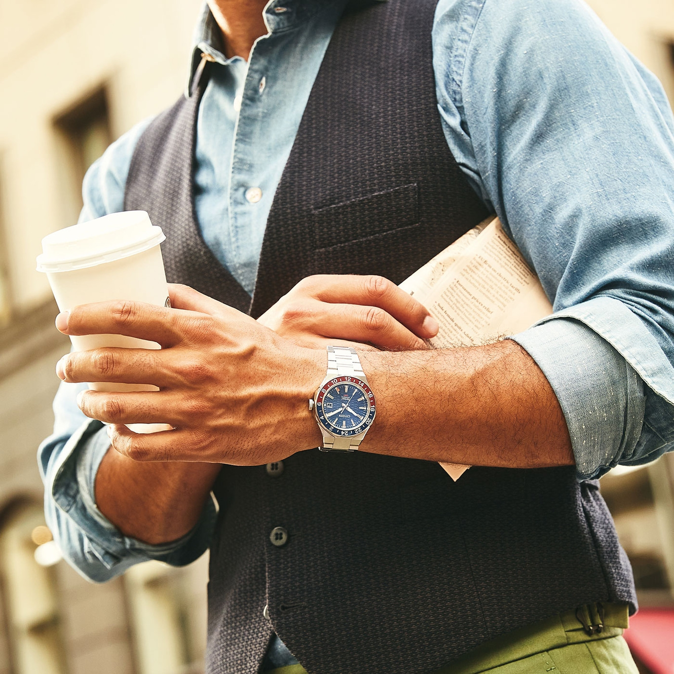 Mann hält Kaffee und Zeitung, trägt eine Uhr und Weste, urbanes Umfeld.