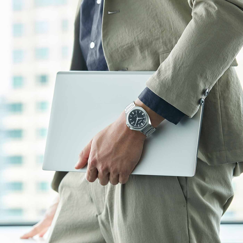 Person hält Laptop, trägt Uhr und Anzug, in der Nähe eines Fensters.