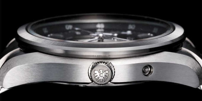 This close-up side view of a watch highlights its brushed metal case and polished bezel, with a textured crown featuring an emblem. The subtle curves and fine finishing are prominent against a dark background, emphasizing its robust yet elegant design.