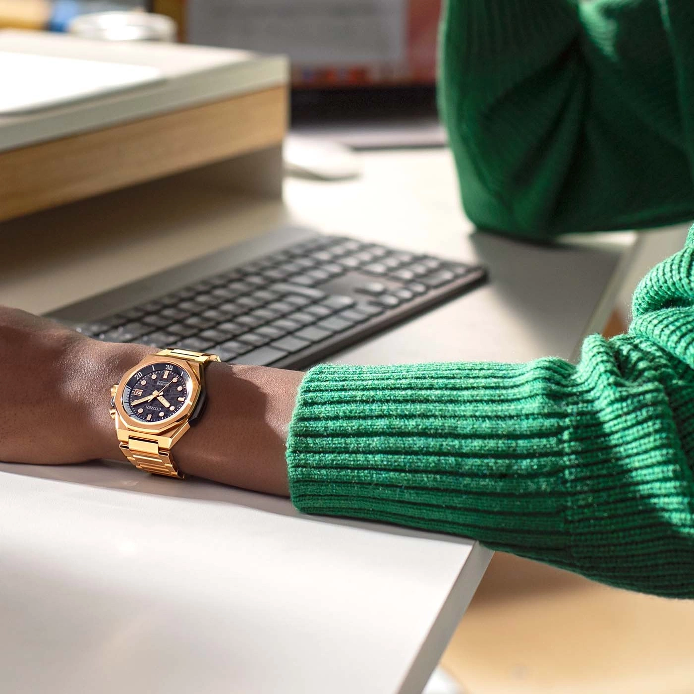 Goldene Uhr am Handgelenk, grüner Pullover, Tastatur, weißer Schreibtisch, Büroumgebung.
