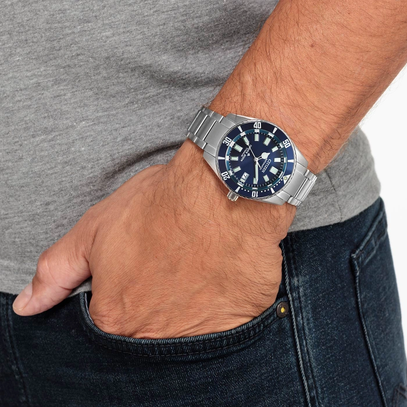 Mann mit Citizen-Uhr, blaues Zifferblatt, Edelstahlarmband, Jeans und graues Hemd.