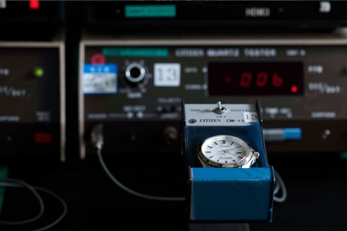 This image shows a silver-toned watch with a white dial placed in a blue testing device, likely a 'CITIZEN CM-10' quartz tester. In the background, various electronic testing equipment with lights and dials are visible, indicating precision measurement.