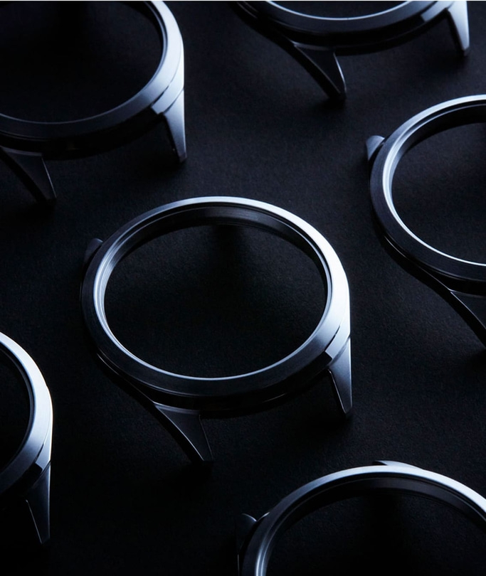 This image shows a collection of polished watch cases arranged on a dark surface, creating a pattern of reflections. Each case is circular with distinct lugs, highlighting their sleek design and the precision involved in their manufacturing.