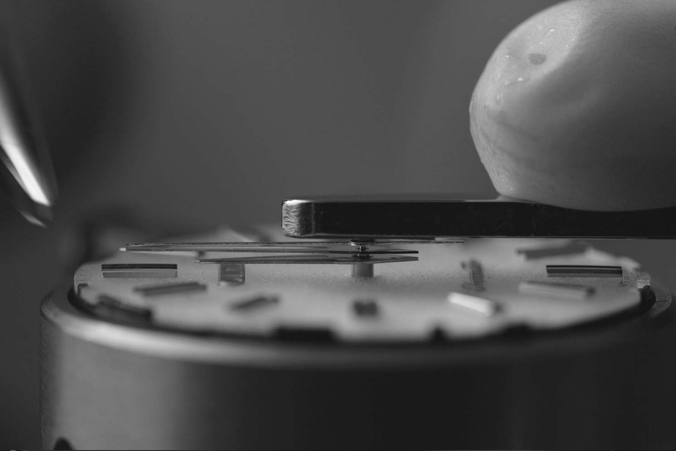 This black and white close-up shows watchmaker's tools precisely working on a watch dial. An instrument positions a component over minute markers, highlighting the delicate, precise nature of watch assembly.