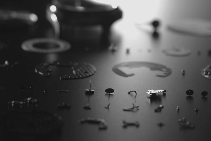 This black and white image displays numerous small, intricate watch components laid out on a dark, reflective surface. The shallow depth of field keeps some parts sharp, highlighting the meticulous detail and precision in watchmaking.