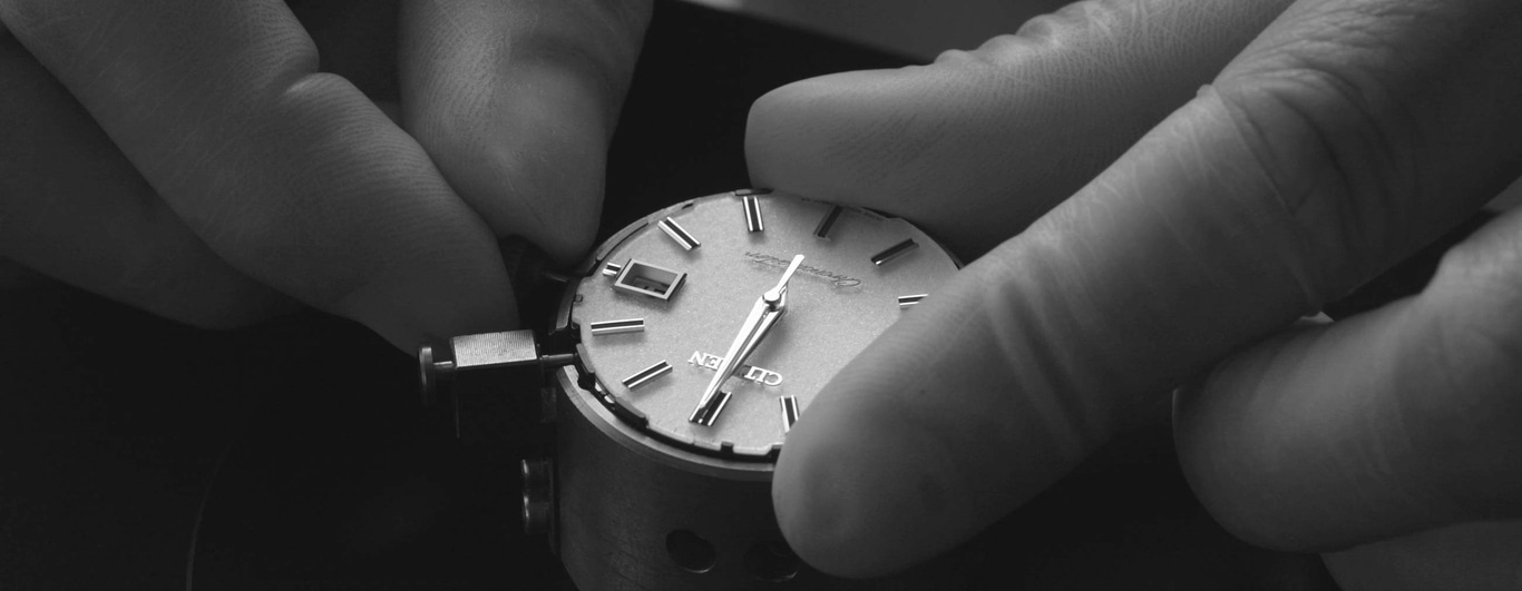This black and white close-up shows a watchmaker's hands precisely working on a watch movement and dial. The hands are positioning or adjusting components, emphasizing the intricate and delicate craftsmanship.