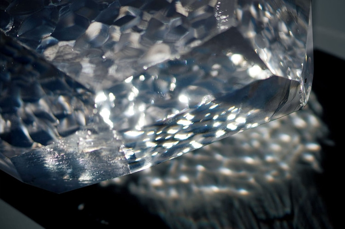 This close-up shows a faceted, clear crystal refracting light. Bright highlights scatter across its textured surface and cast dappled reflections onto a dark background, creating a dazzling, abstract effect.