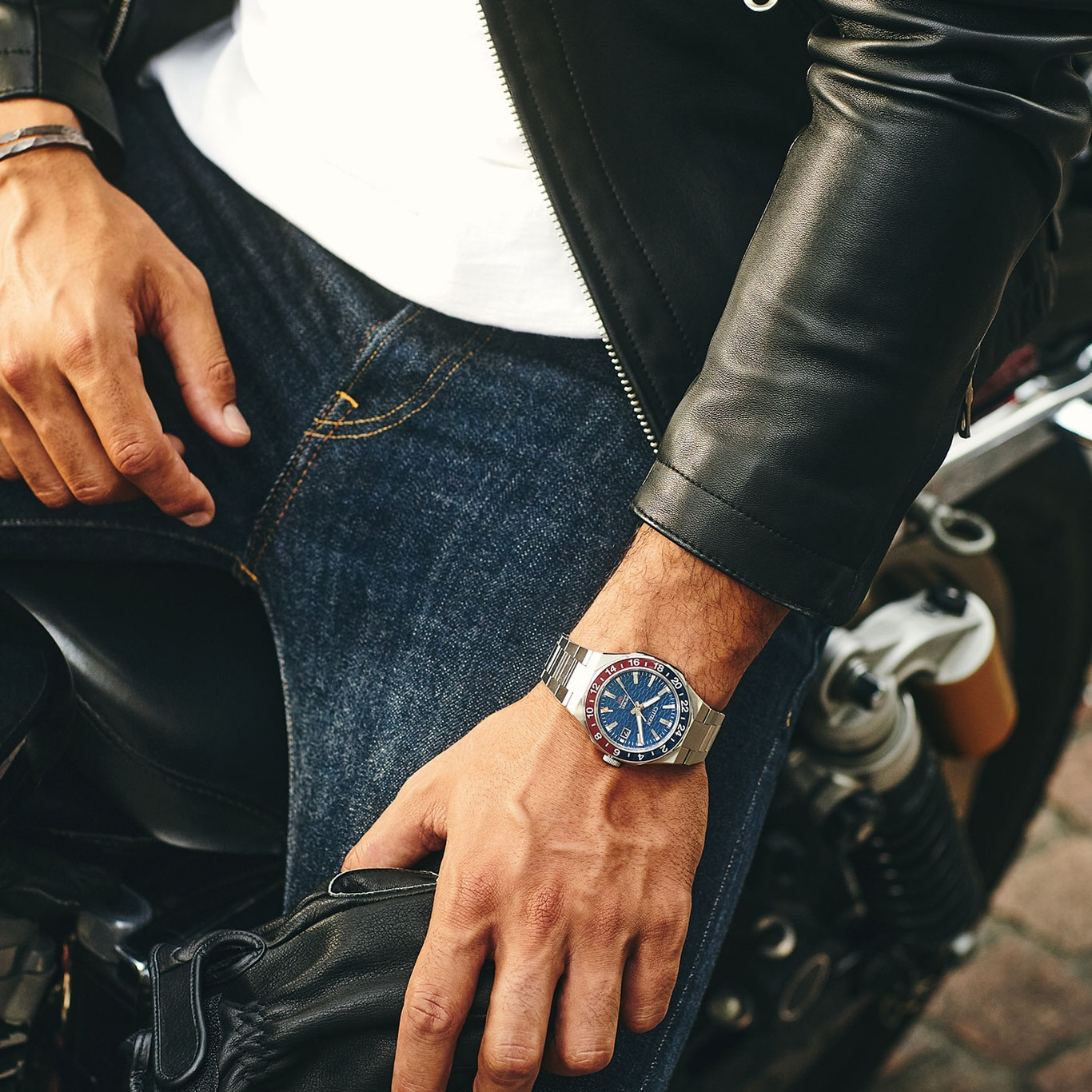Mann mit Uhr, Lederjacke und Jeans auf einem Motorrad.