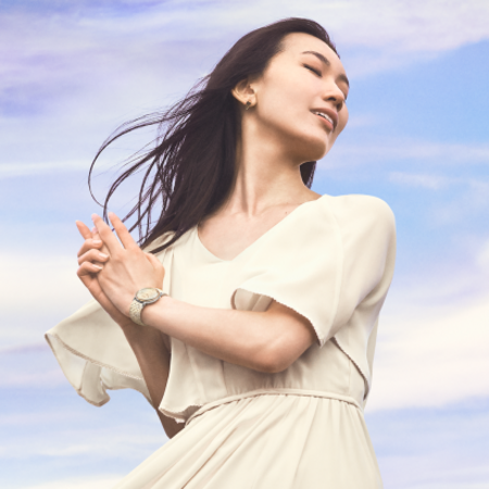 A woman in a flowing white dress stands with her hands clasped against a backdrop of soft blue sky, exuding serenity.
