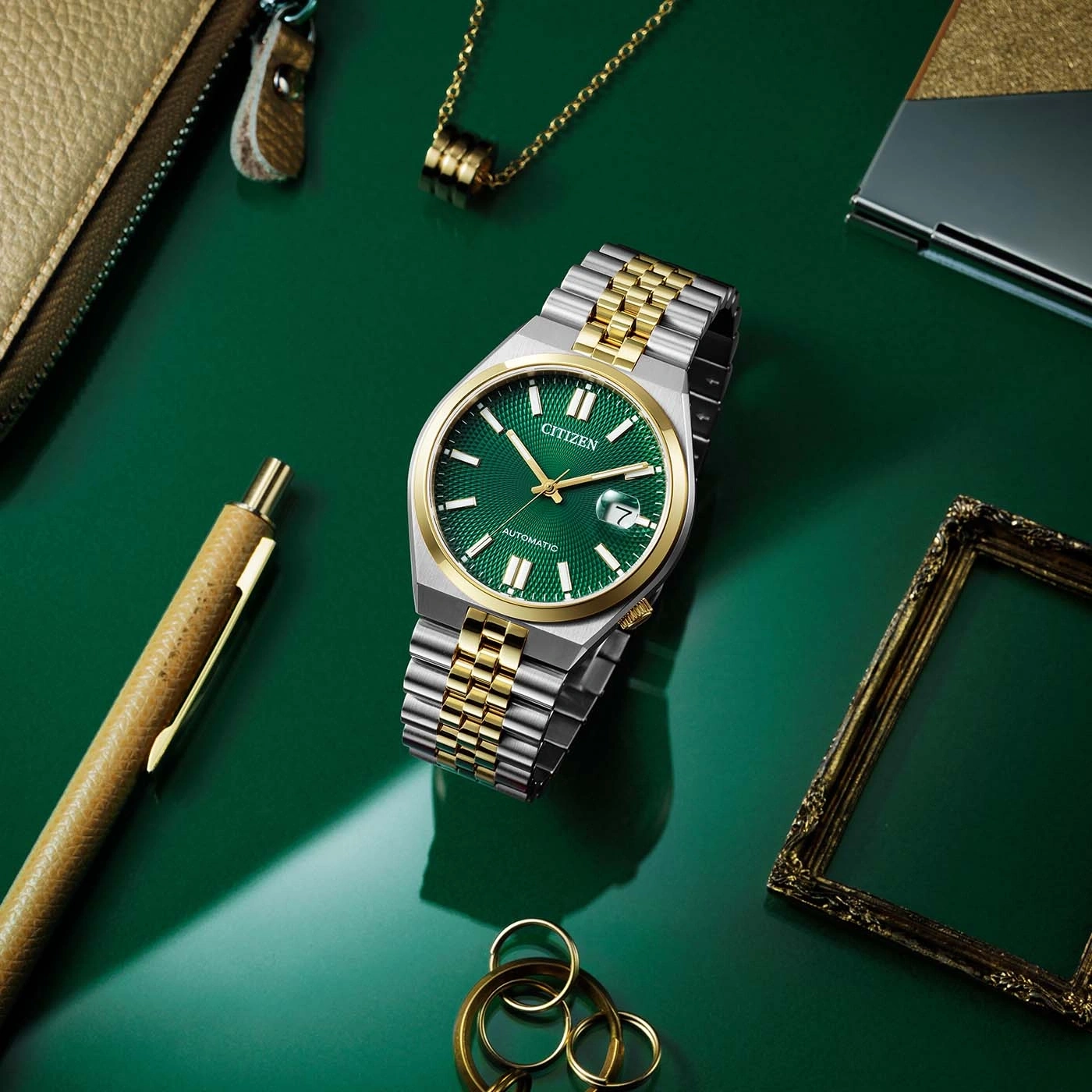 Citizen-Uhr mit grünem Zifferblatt, Gold- und Silberarmband, auf grüner Oberfläche mit Zubehör.