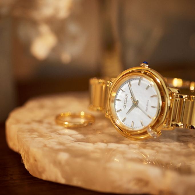 A luxurious close-up of a golden Citizen watch and a matching ring resting on a marble surface, bathed in warm, soft light.
