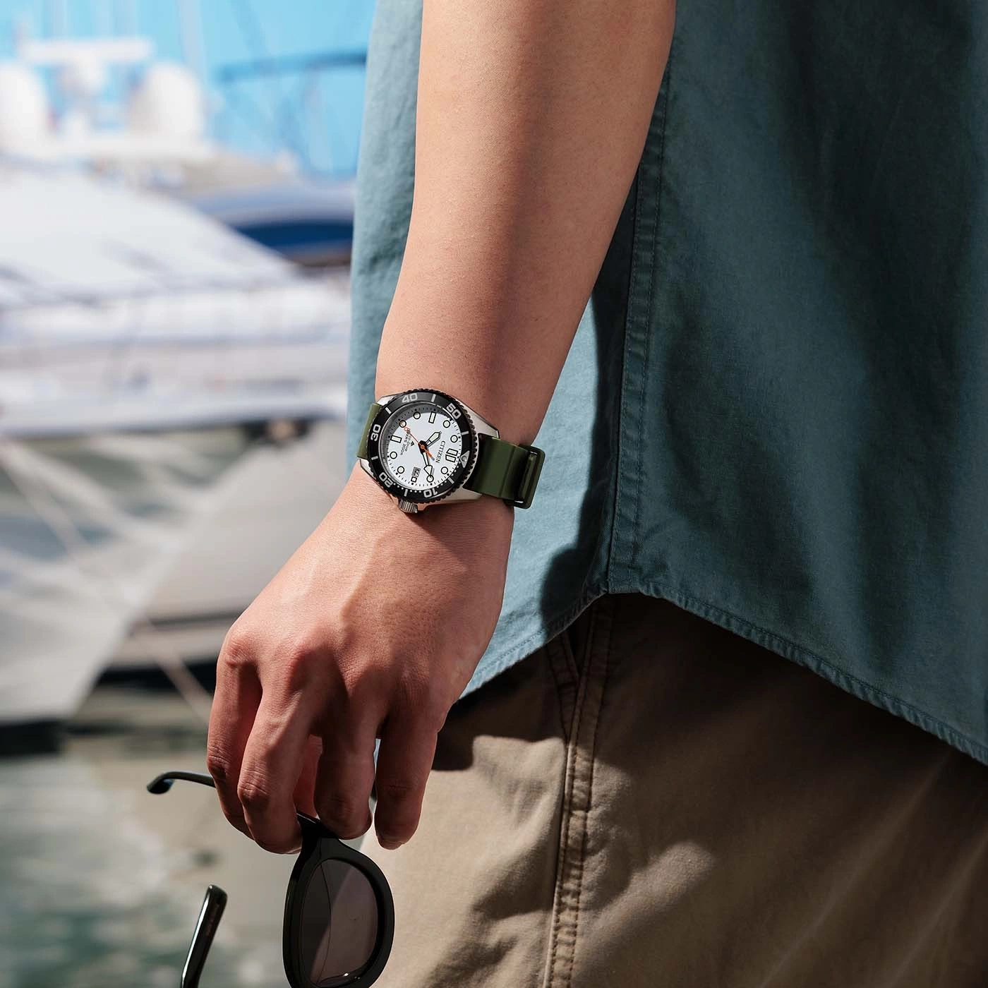 Mann mit Uhr und Sonnenbrille am Wasser, Boote im Hintergrund.