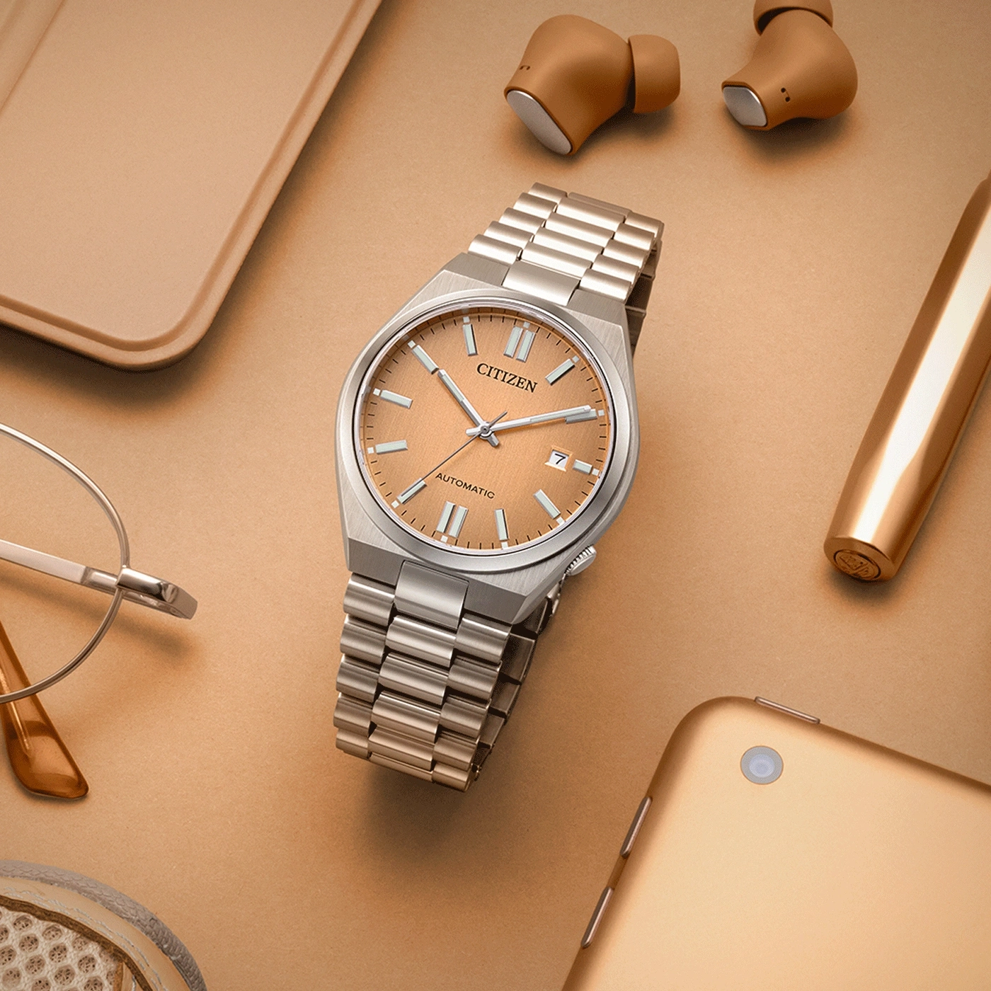 Citizen-Uhr, Ohrhörer, Stift, Brille und Tablet auf goldener Oberfläche.