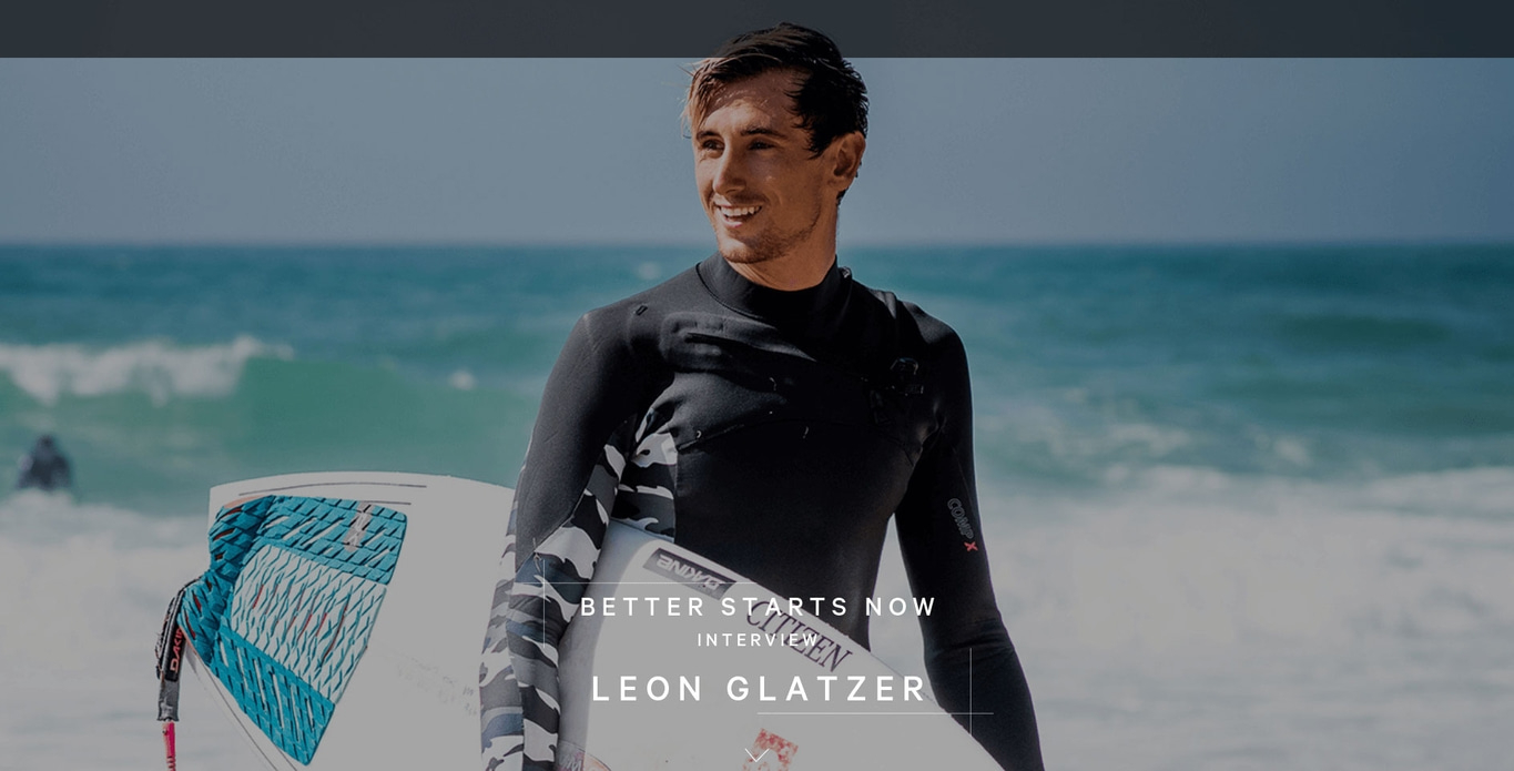 Mann im Neoprenanzug mit Surfbrett am Strand, lächelnd, Interview mit Leon Glatzer