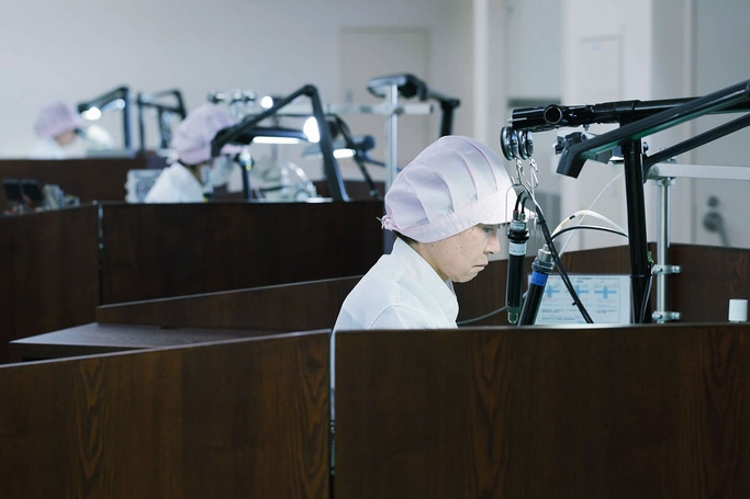 This image shows workers, wearing caps, focused intently at individual workstations. Each station has specialized equipment, suggesting precision assembly or detailed examination in a controlled environment like a watchmaking factory.